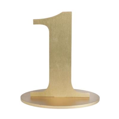 En bois coloris or, le chiffre 1 posé sur son  support à utiliser comme marque table ou en déco de table anniversaire.